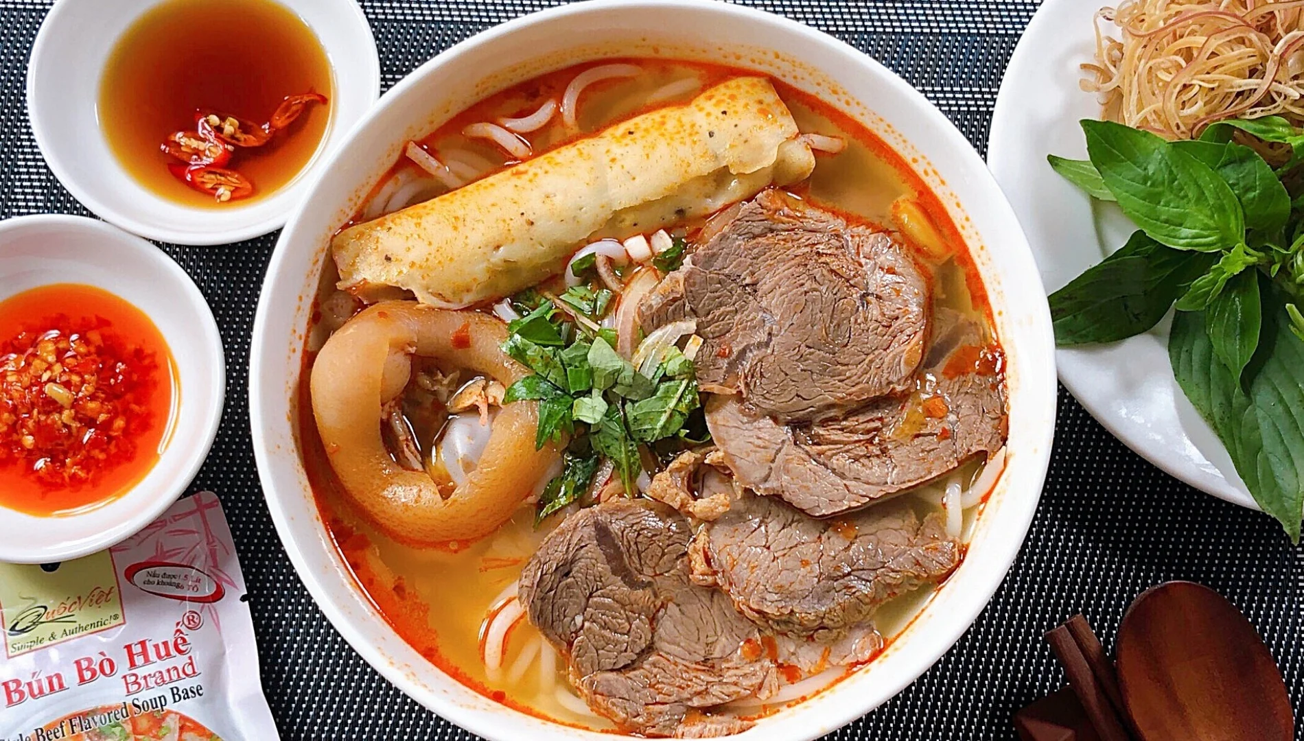 A bowl of Bun Bo Hue (Hue Beef Noodle Soup) featuring a rich broth, beef, pig's trotter, and fresh herbs.