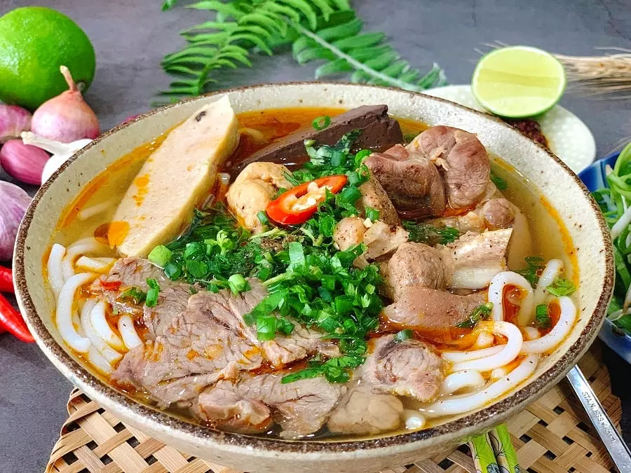 A steaming bowl of Hue Beef Noodle Soup with beef slices, herbs, and spices.