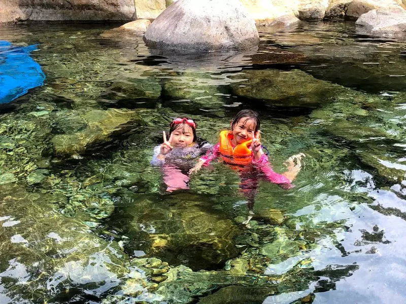 Visitors swimming in the crystal-clear, emerald waters of Elephant Springs, with colorful pebbles visible at the bottom, surrounded by lush nature.