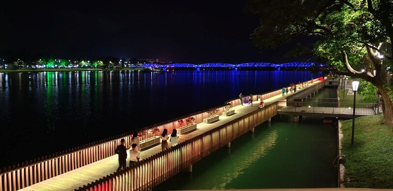 Hue Lim Wood Bridge lit up at night with colorful lights, casting reflections on the Huong River.